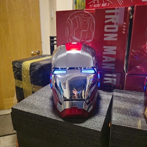 Puede incluir: Un casco de Iron Man rojo y plateado con luces azules. El casco está sobre una superficie negra y el texto "IRON MAN" es visible en la caja detrás de él.