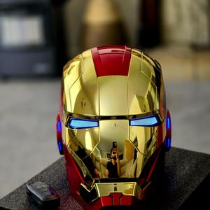 May include: A gold and red Iron Man helmet with blue LED lights. The helmet is made of plastic and has a glossy finish.