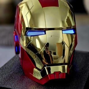 May include: A gold and red Iron Man helmet with blue LED lights for the eyes. The helmet is made of a shiny, metallic material.