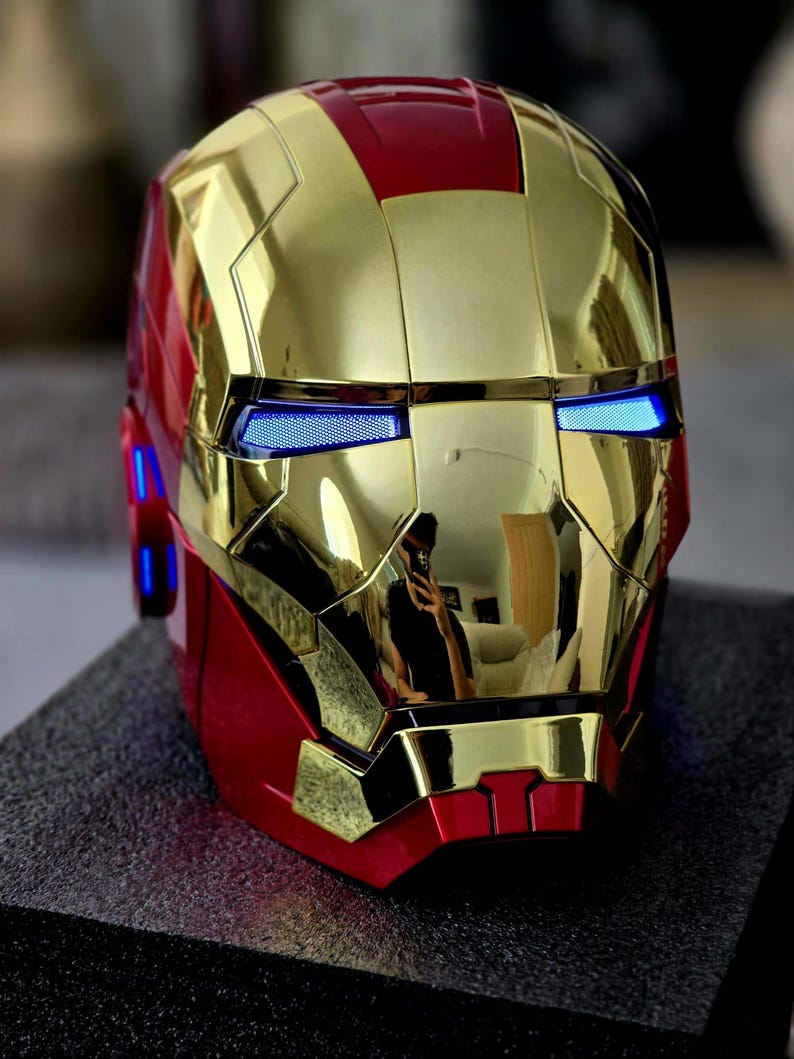 May include: A gold and red Iron Man helmet with blue LED lights for the eyes. The helmet is made of a shiny, metallic material.