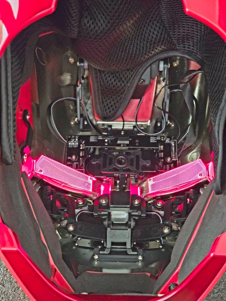 May include: An internal view of a red helmet with a black mesh lining. The helmet features a black and silver mechanism with two pink lights on either side.