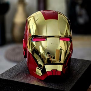 May include: A gold and red Iron Man helmet with a red visor. The helmet is made of plastic and has a shiny finish.