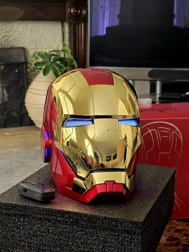 May include: A red and gold Iron Man helmet with glowing blue eyes. The helmet is on a black surface and has a remote control next to it.