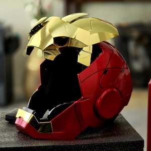 May include: A red and gold Iron Man helmet replica. The helmet is made of plastic and has a detailed design. The helmet is on a black surface.