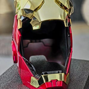 May include: A red and gold helmet with a black interior. The helmet has a faceplate that is partially open.