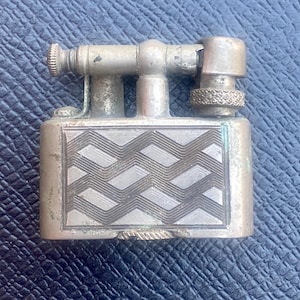 May include: A silver-toned lighter with a geometric pattern on the front. The lighter has a lever on the side and a small knob on the top.