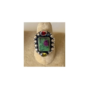 May include: A silver ring with a rectangular green stone with a red center. The ring is accented with a red and yellow stone, and surrounded by small silver beads. The ring is displayed on a beige surface.