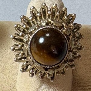 May include: A vintage ring featuring a large, round, polished tiger's eye stone. The stone is set in a gold-toned metal setting with a sunburst design, including leaf and bead details. The ring is displayed on a neutral-toned surface.