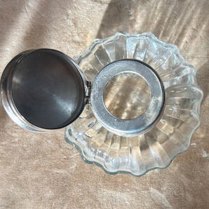 Puede incluir: Un tintero de vidrio transparente con tapa y borde de metal plateado. El tintero tiene una abertura circular y una base acanalada y festoneada. La tapa est&aacute; abierta, revelando el interior del tintero.