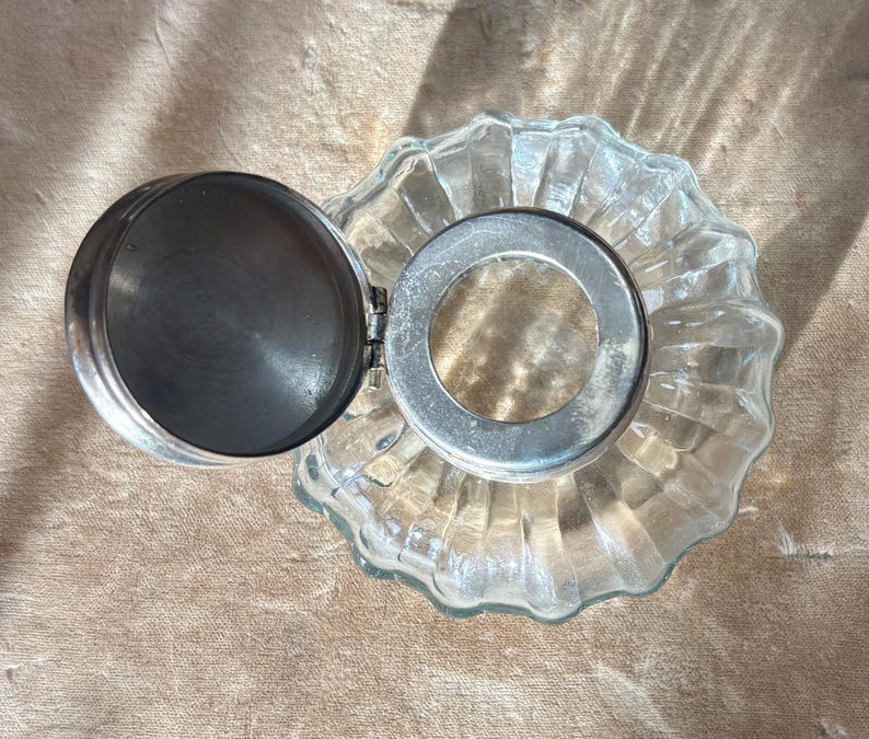 Puede incluir: Un tintero de vidrio transparente con tapa y borde de metal plateado. El tintero tiene una abertura circular y una base acanalada y festoneada. La tapa est&aacute; abierta, revelando el interior del tintero.