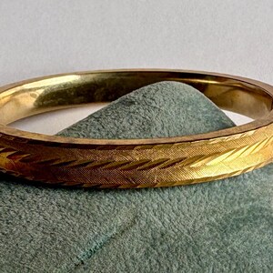 May include: A gold-colored hinged bangle bracelet with a textured, etched design. The bracelet is resting on a folded, teal-colored fabric. The bracelet's surface has a repeating pattern of diagonal lines.