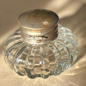 Puede incluir: Un tintero de vidrio transparente con un cuerpo redondeado y acanalado y una tapa met&aacute;lica con bisagras y deslustrada. El tintero tiene una est&eacute;tica vintage, mostrando un dise&ntilde;o cl&aacute;sico y funcional. La tapa est&aacute; cerrada.
