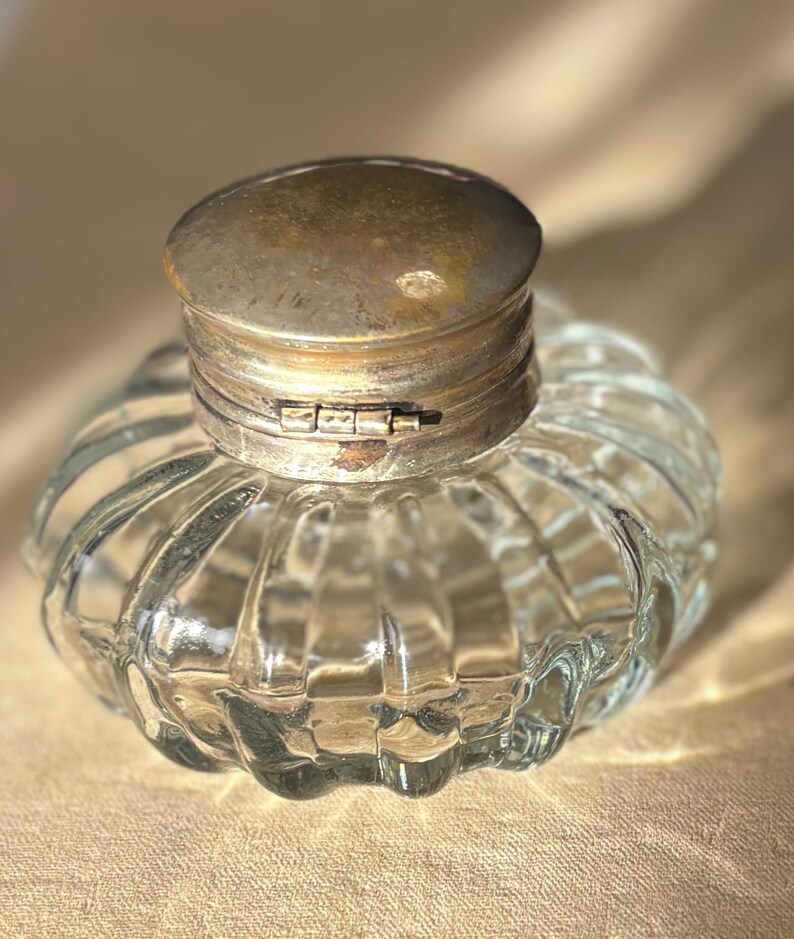 Puede incluir: Un tintero de vidrio transparente con un cuerpo redondeado y acanalado y una tapa met&aacute;lica con bisagras y deslustrada. El tintero tiene una est&eacute;tica vintage, mostrando un dise&ntilde;o cl&aacute;sico y funcional. La tapa est&aacute; cerrada.