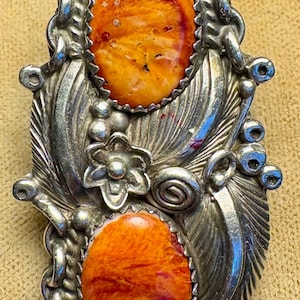 May include: A silver-toned, ornate ring featuring two oval-shaped, orange-red stones. The ring has a detailed design with leaf and floral accents, and a chain-like border. The stones have a textured appearance.