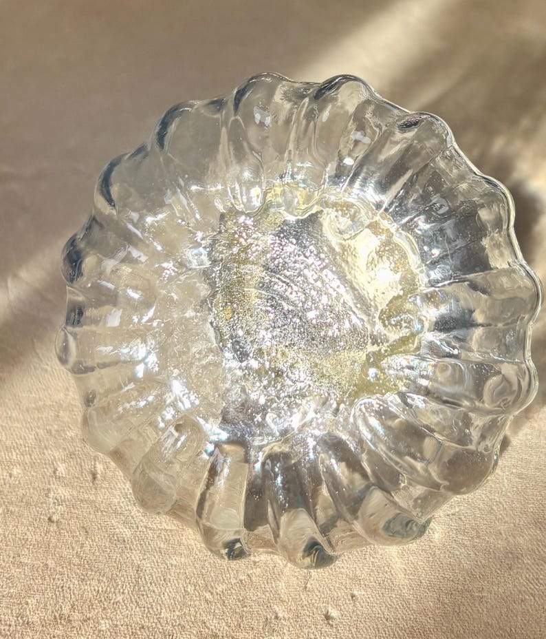 Puede incluir: Un cuenco decorativo de vidrio transparente con un exterior texturizado y acanalado. El centro del cuenco tiene un acabado met&aacute;lico plateado y dorado brillante. El cuenco es redondo y se asienta sobre una superficie de color neutro.