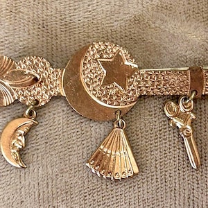 May include: A gold-toned brooch with a crescent moon, star, and fan design. The brooch has a textured surface and features dangling charms, including a crescent moon, a fan, and a pair of scissors. The brooch is likely vintage or antique.