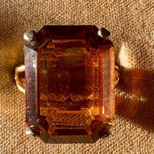 May include: A rectangular, amber-colored gemstone ring with a gold band. The gemstone has a faceted cut, and small silver-colored prongs secure it to the band. The ring is set against a textured, tan background.