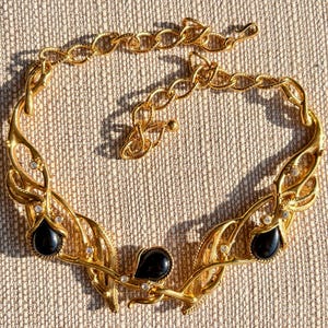 May include: A gold-toned necklace with a leaf-like design, featuring three black teardrop-shaped stones and small clear crystals. The necklace has an adjustable chain and is displayed on a textured beige surface.