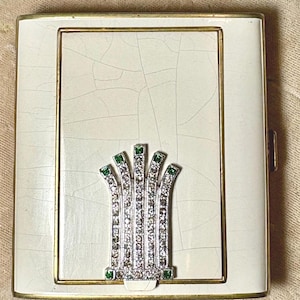 May include: A vintage compact mirror with a white and gold frame. The center features a decorative silver design with clear and green rhinestones. The compact has a gold clasp and a cracked, textured surface.