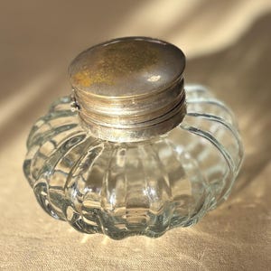 Puede incluir: Un tintero de vidrio transparente con un cuerpo redondeado y acanalado y una tapa de metal. La tapa es de color plateado deslustrado. El tintero est&aacute; vac&iacute;o y se encuentra sobre una superficie de tono neutro. Dise&ntilde;o vintage.