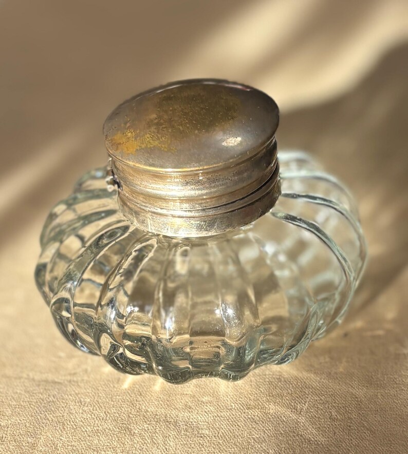 Puede incluir: Un tintero de vidrio transparente con un cuerpo redondeado y acanalado y una tapa de metal. La tapa es de color plateado deslustrado. El tintero est&aacute; vac&iacute;o y se encuentra sobre una superficie de tono neutro. Dise&ntilde;o vintage.
