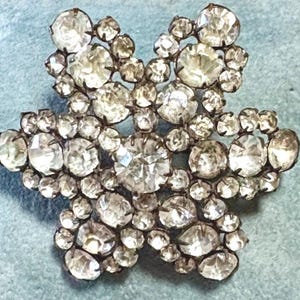 May include: A vintage-style brooch in a floral design, featuring numerous clear, faceted rhinestones set in a silver-tone metal. The brooch sparkles with light, showcasing its intricate details and craftsmanship.