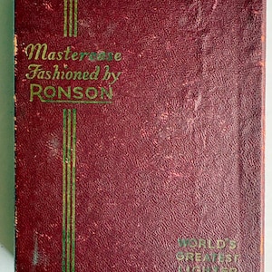 May include: A vintage burgundy Mastercase by Ronson. The cover features the text "Mastercase Fashioned by Ronson" in gold lettering, with vertical green stripes. The bottom right corner reads "World's Greatest Lighter".