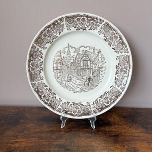 May include: A round ceramic plate with a cream-coloured base and a dark brown transferware pattern. The rim features a detailed floral border, while the centre depicts a street scene with timber-framed buildings, a church spire, and figures.