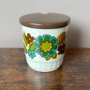 May include: A white ceramic canister with a brown lid featuring a floral pattern in shades of green, blue, and brown. The cylindrical container has a ribbed base and a small notch in the lid for a spoon, showcasing a colourful botanical design.