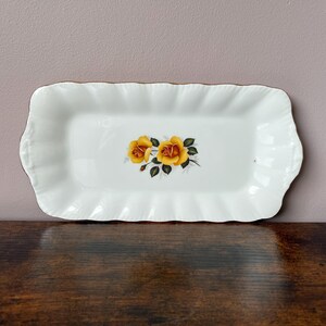 May include: A rectangular white porcelain serving tray with scalloped edges and a gold-trimmed rim. The centre of the tray features a detailed illustration of two vibrant yellow roses with green leaves and stems against a plain white background.