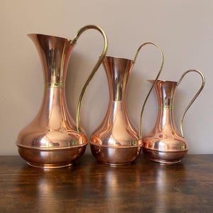 May include: A set of three graduated copper jugs with polished surfaces, each featuring a slender neck, a wide rounded base, and a curved brass handle. The jugs are arranged in a row from largest to smallest, showcasing a metallic finish.