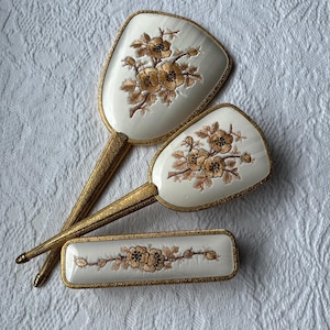 May include: A vanity set consisting of two hand mirrors and a rectangular brush, each featuring an off-white surface with a gold-toned floral design and a textured gold-coloured metal frame and handle, arranged in a layered composition.
