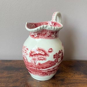 May include: A cream-coloured ceramic jug featuring a red transferware pattern of a landscape with trees, buildings, and hills. The jug has a scalloped rim, a curved handle, and a floral border design around the top edge of the vessel.