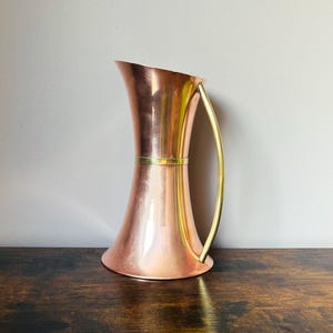 May include: A polished copper pitcher with a flared top and base, featuring a smooth, hourglass-shaped body. A thin, horizontal brass band encircles the centre, and a curved, slender brass handle is attached to the side of the vessel.