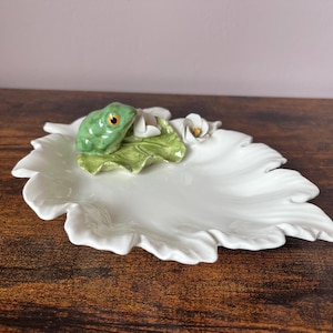 May include: A white ceramic leaf-shaped dish with a green frog figurine resting on a green leaf. The frog has orange eyes. Two small white flowers are also on the dish. The dish is on a wooden surface.