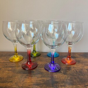 May include: A set of five clear glass goblets with rounded bowls and stems featuring distinct colour gradients. The stems and bases are coloured in yellow, red, green, blue, and orange, creating a vibrant collection of drinking glasses.