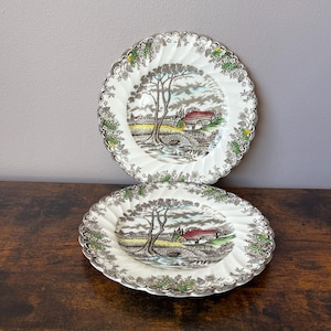 May include: Two round ceramic plates featuring a scalloped edge and a detailed transferware scene of a rural landscape with a stone bridge, a cottage with a red roof, and a large tree. The border is decorated with green and yellow floral motifs.