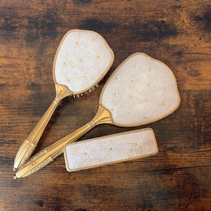 May include: A vanity set consisting of a hairbrush, a hand mirror, and a rectangular brush, all featuring gold-toned metal handles with ribbed detailing and off-white fabric inserts decorated with an embroidered floral and swirling vine pattern.