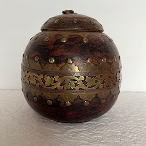 May include: A round wooden box with a lid, decorated with brass accents and intricate floral designs. The box has a dark brown wood finish and is accented with brass studs.