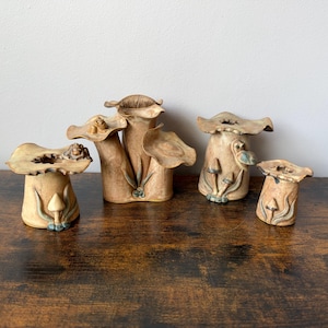 May include: A set of four tan ceramic vases with organic, mushroom-inspired shapes. Each vase features a wide, wavy rim and sculpted mushroom details at the base, with subtle blue-green accents on the stems and caps of the small mushrooms.