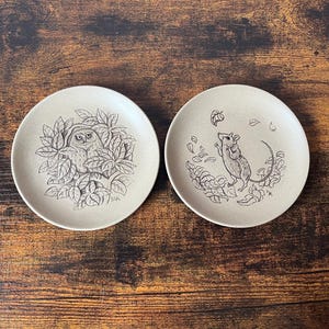 May include: Two round, beige ceramic plates featuring black line art illustrations. The left plate depicts an owl nestled within dense foliage. The right plate shows a mouse standing among scattered leaves. Both plates have a matte, speckled finish.
