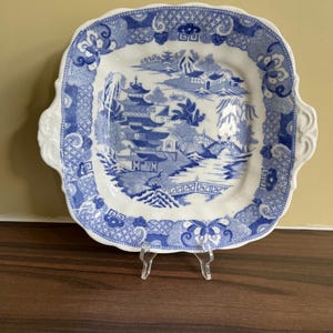 May include: Square, white and blue decorative plate with a scenic landscape design. The plate features a blue and white colour scheme with a detailed illustration of a bridge, buildings, and trees. The plate has a scalloped edge and two handles.