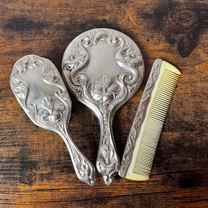 May include: A silver-toned vanity set consisting of a hand mirror, a hairbrush, and a comb. Each piece features an ornate, raised floral relief pattern. The comb has a cream-coloured plastic base with a metallic handle featuring the same floral design.