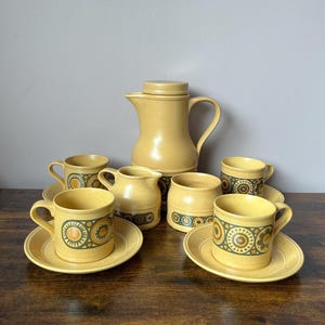 May include: A yellow ceramic coffee set featuring a lidded pitcher, a creamer, a sugar bowl, and four cups with matching saucers. Each piece displays a speckled finish and a decorative band of dark green and orange circular geometric patterns.