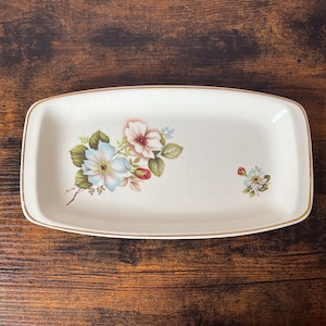May include: A rectangular off-white ceramic serving tray with rounded corners and a thin gold rim. The centre features a floral illustration of blue and white flowers with green leaves, accompanied by a smaller matching floral motif to the right.