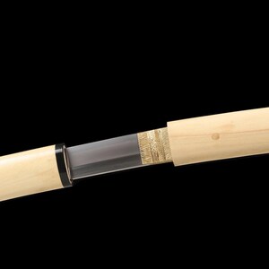 May include: A Japanese katana sword with a light-colored wooden scabbard. The blade is partially visible, with a dark gray color. The hilt features a black ring. The scabbard has a gold-colored decorative element near the blade.