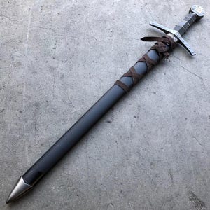 May include: A black sword with a brown leather wrapped handle and a silver pommel. The sword is lying on a gray concrete surface.