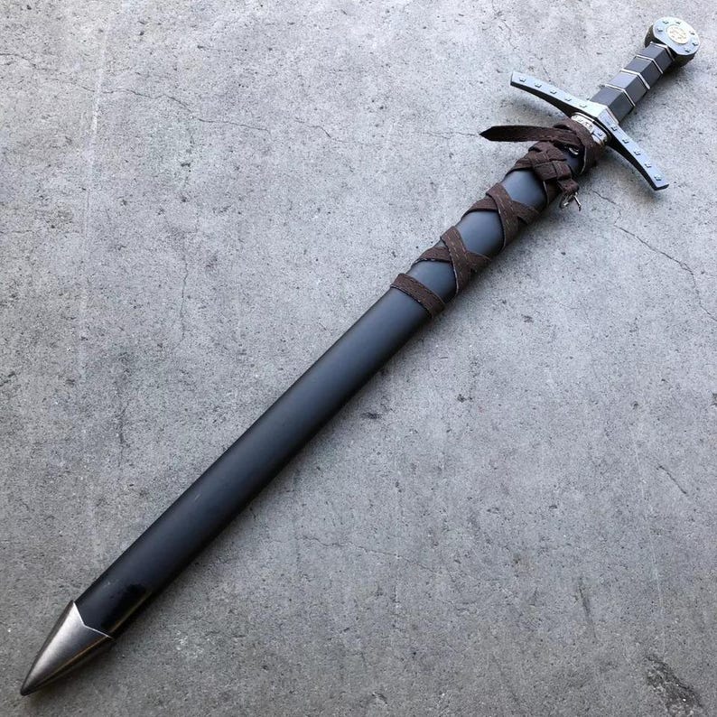May include: A black sword with a brown leather wrapped handle and a silver pommel. The sword is lying on a gray concrete surface.