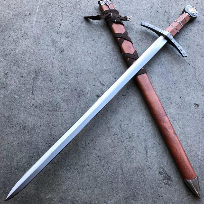 May include: A silver sword with a brown leather wrapped handle and a brown wooden scabbard. The sword is lying on a gray concrete surface.