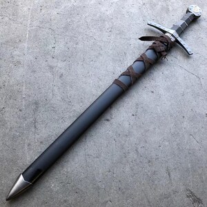 May include: A black sword with a brown leather wrapped handle and a silver pommel. The sword is lying on a gray concrete surface.